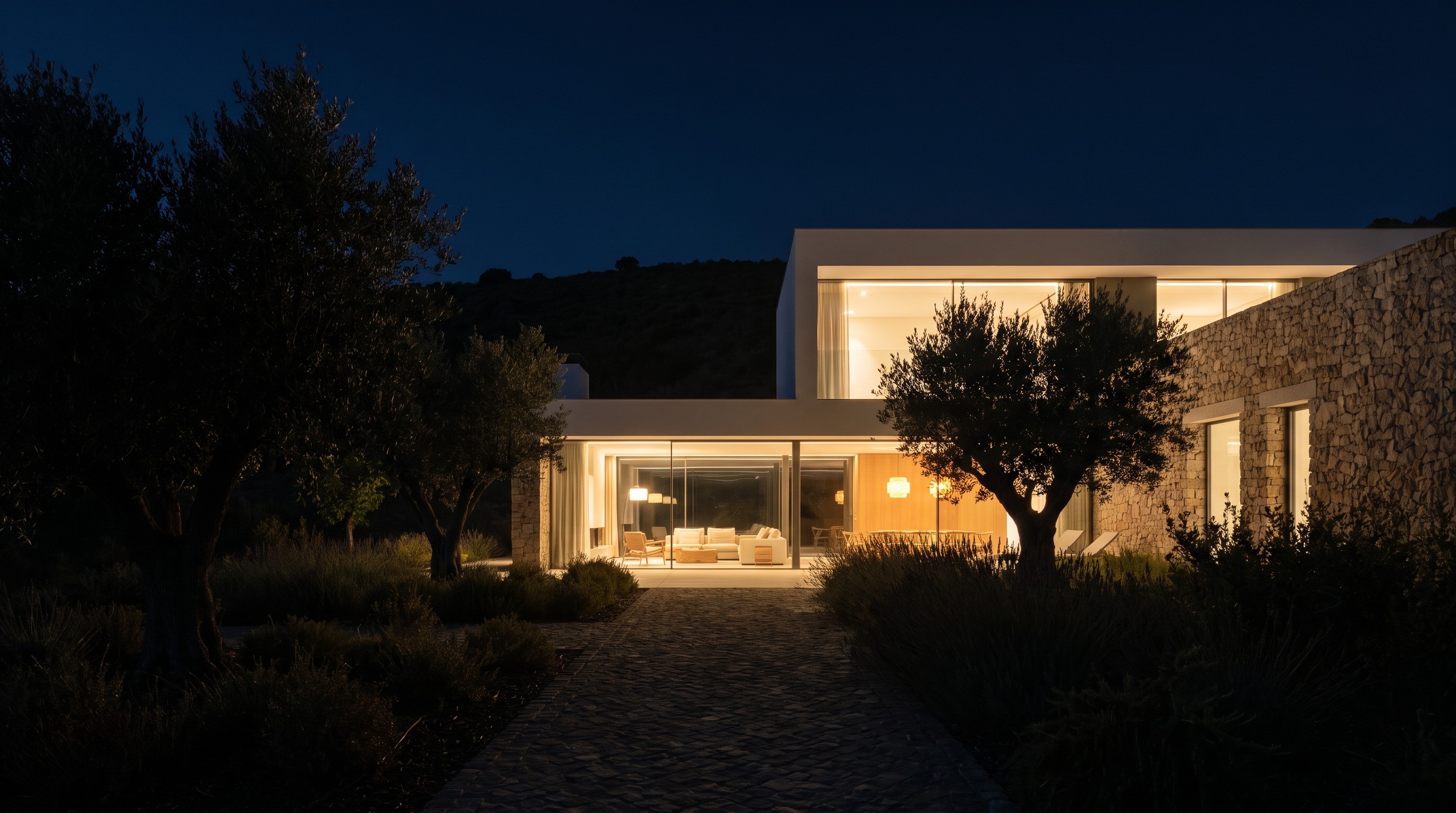 Moradia contemporânea iluminada por dentro, vista do jardim escuro, com a encosta em redor às escuras